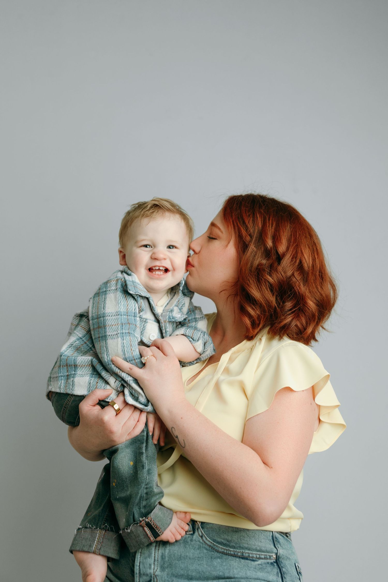 Red-haired mother kissing smiling toddler boy in plaid shirt at the family portrait specializing studio in Victoria BC.