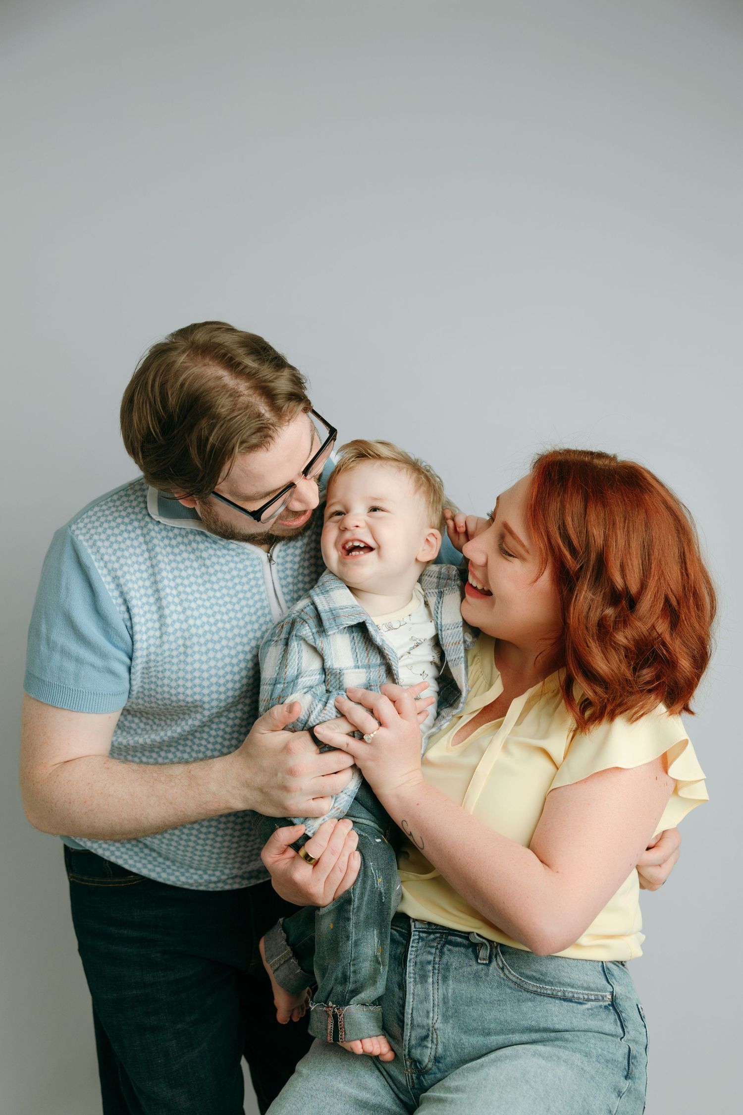 Happy young family with redhead mom, dad in glasses, and laughing toddler posing together at the family portrait studio in Victoia BC