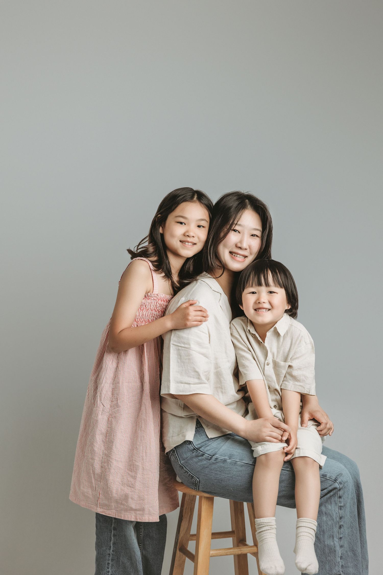 A smiling Asian mother sits on a stool with her two daughters hugging her at the family portrait specializing studio in Victoria BC.