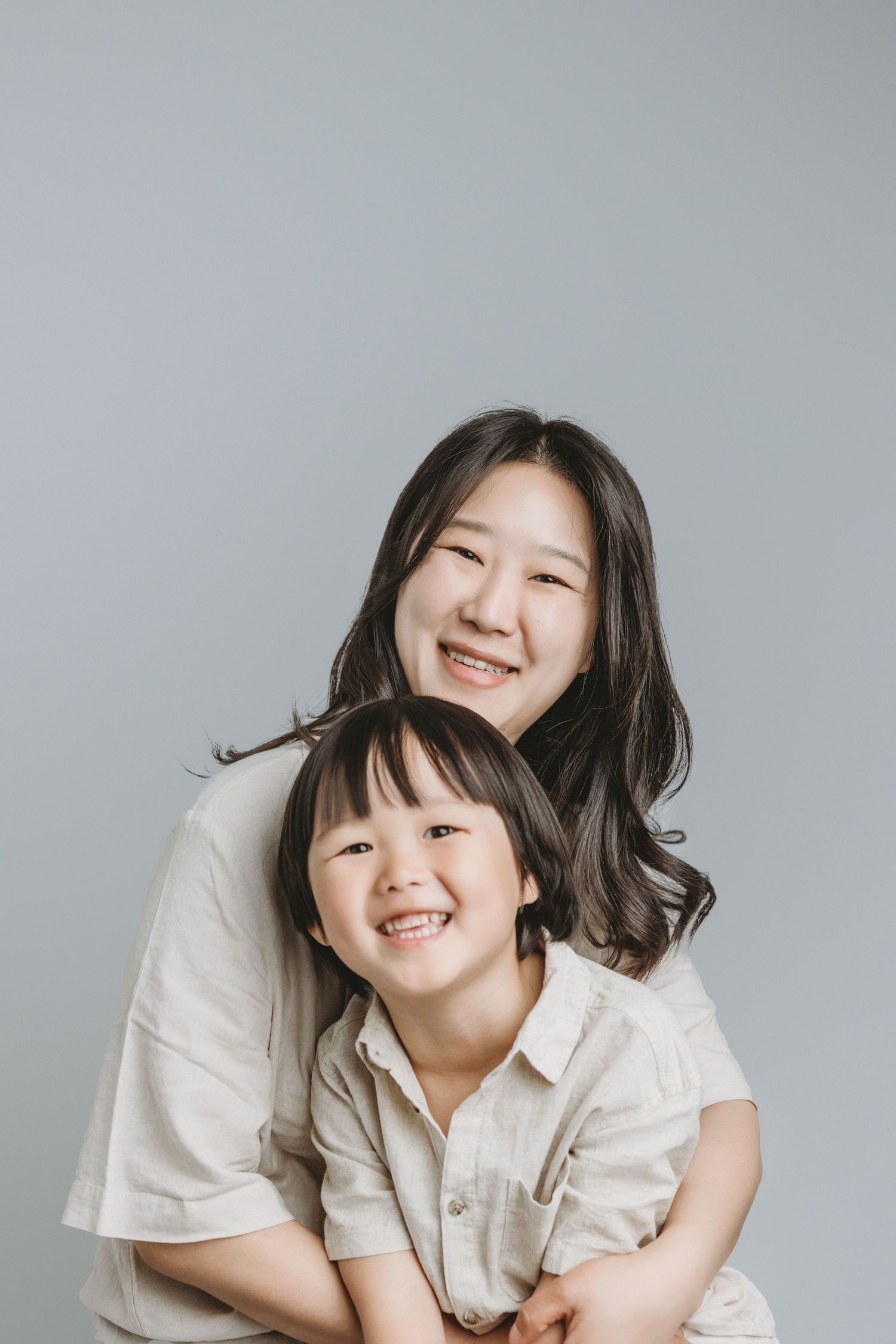 Smiling Asian mother hugging her young child, both wearing white shirts at the family portrait specializing studio in Victoria BC
