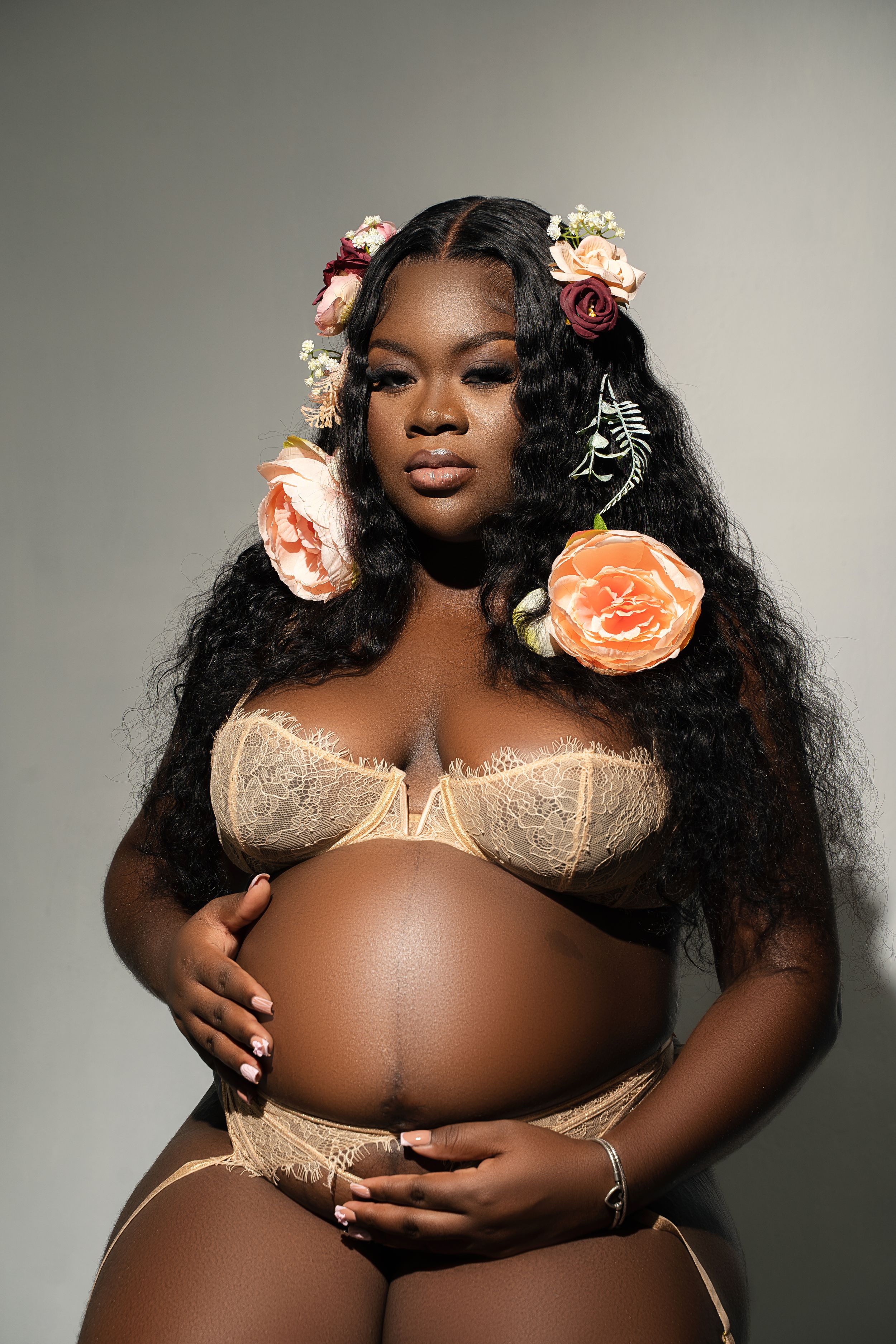 A model poses in beige lingerie with peach roses adorning long wavy black hair against a grey studio backdrop.