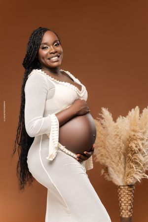 Beautiful maternity photo in white dress with long braids posing next to pampas grass against warm brown backdrop.