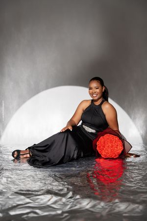 Elegant portrait against silver backdrop featuring black evening gown and vibrant red round hatbox with reflective floor.