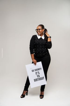 A person in glasses and black polka dot top poses against gray background holding a sign about a birthday celebration.