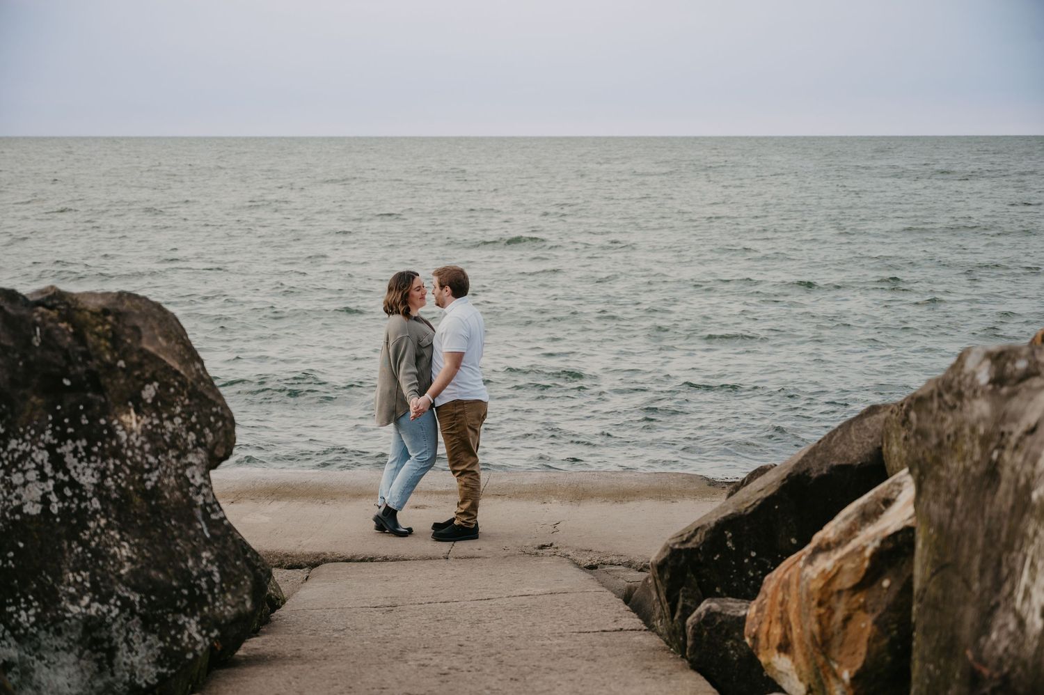 Couple holding hands and kissing on a rocky beach pier with ocean waves behind them.