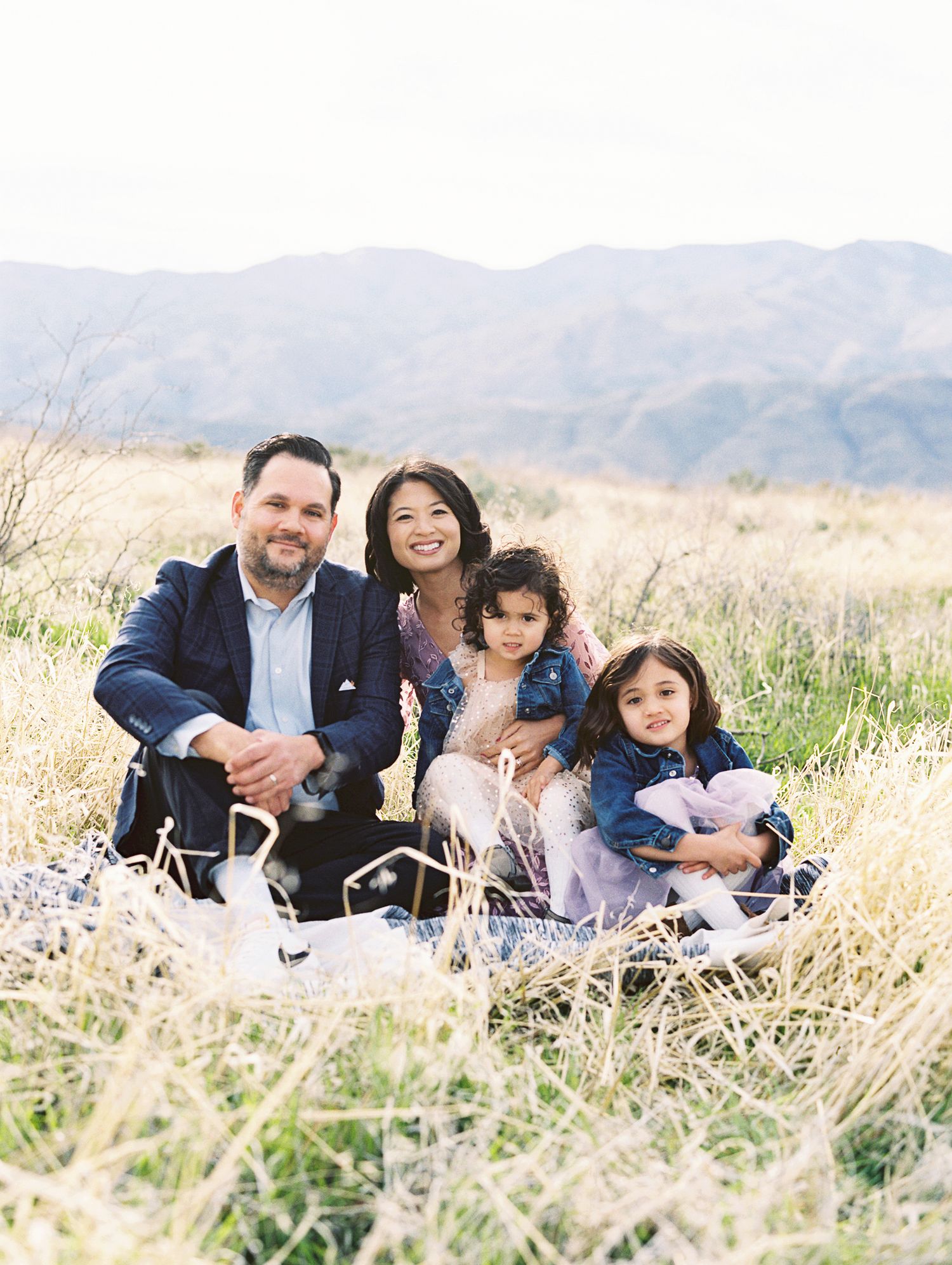 Family of four smiling together in a grassy field with mountain backdrop during an outdoor photo session.