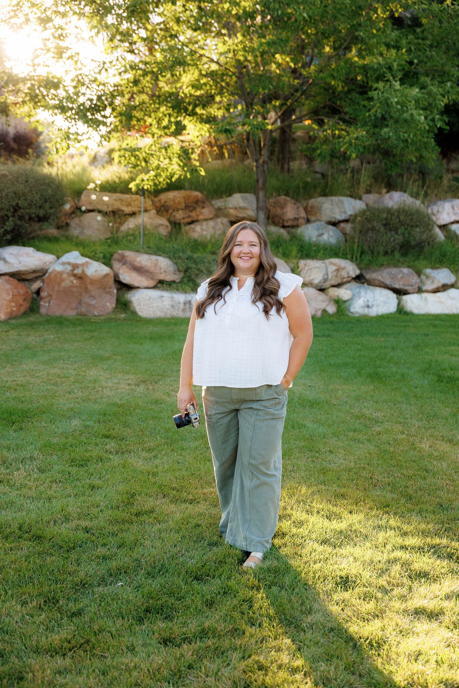 Woman photographer smiling while holding a camera outdoors in a green field.