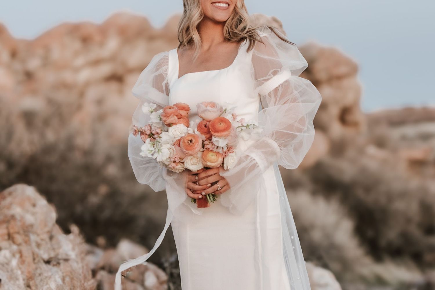 utah Bride in flowing white dress poses with coral flowers in desert setting.