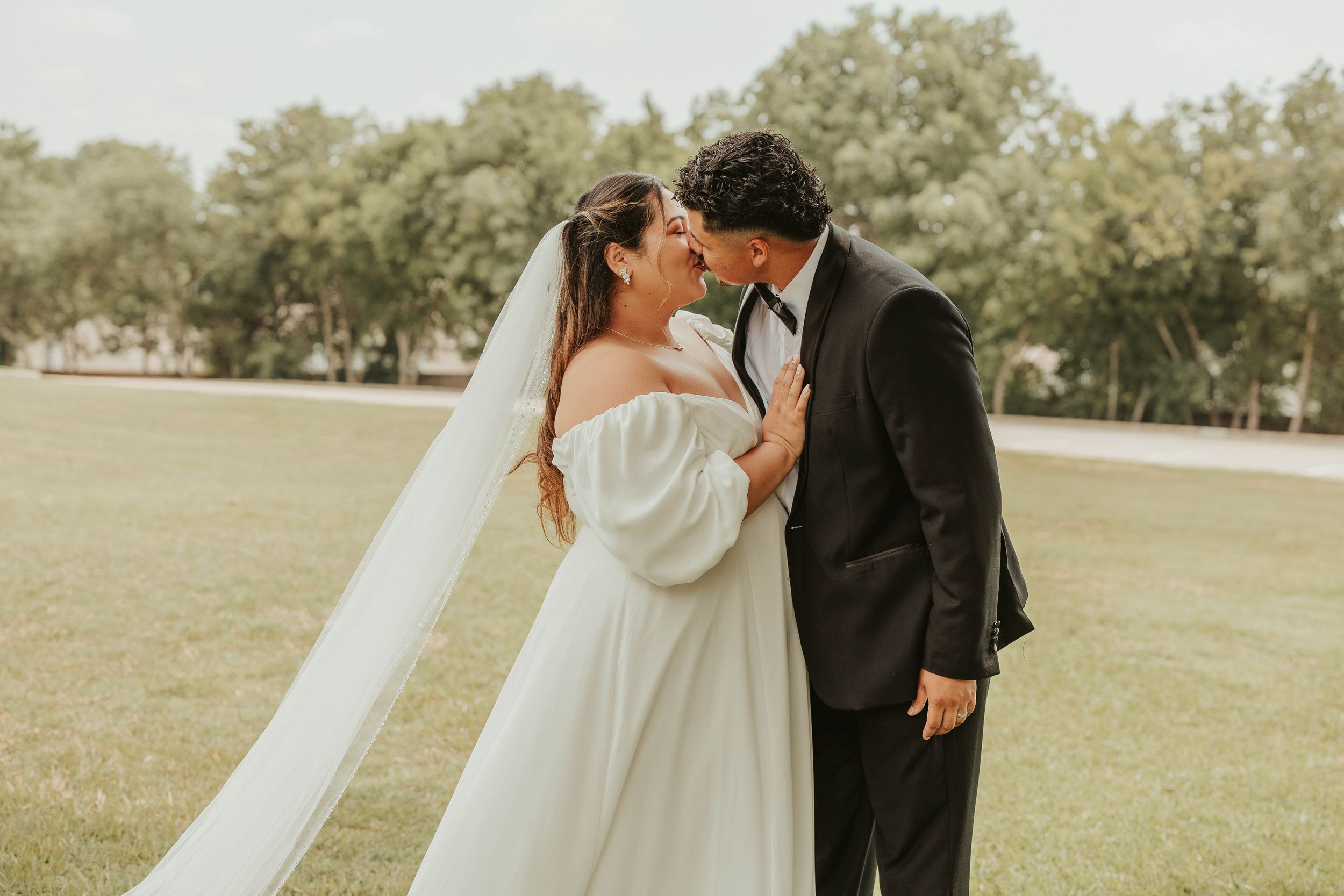 Bride in a flowing white dress and groom in black suit sharing a romantic moment outdoors.