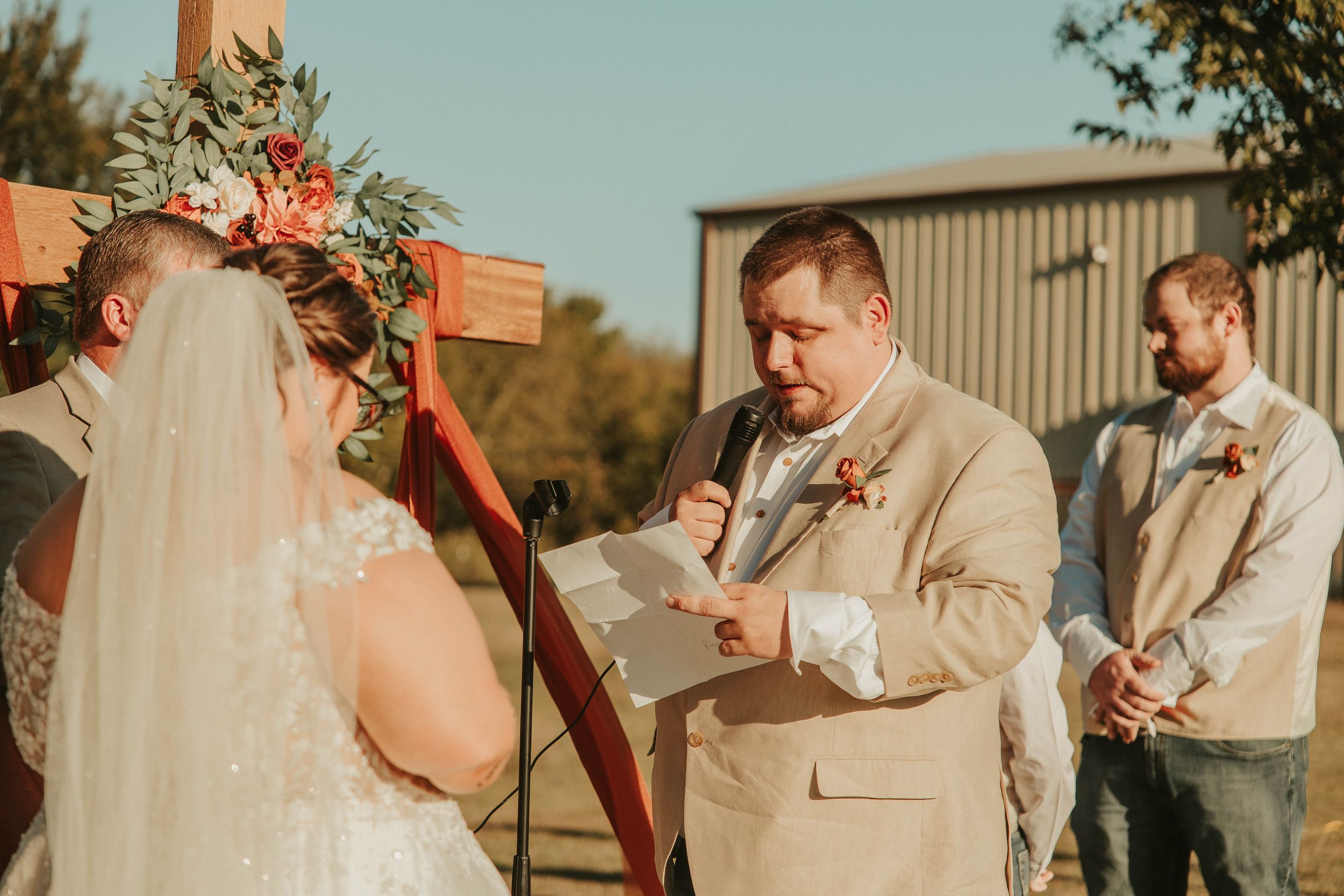 Wedding ceremony taking place outdoors with wooden arch and floral decorations.