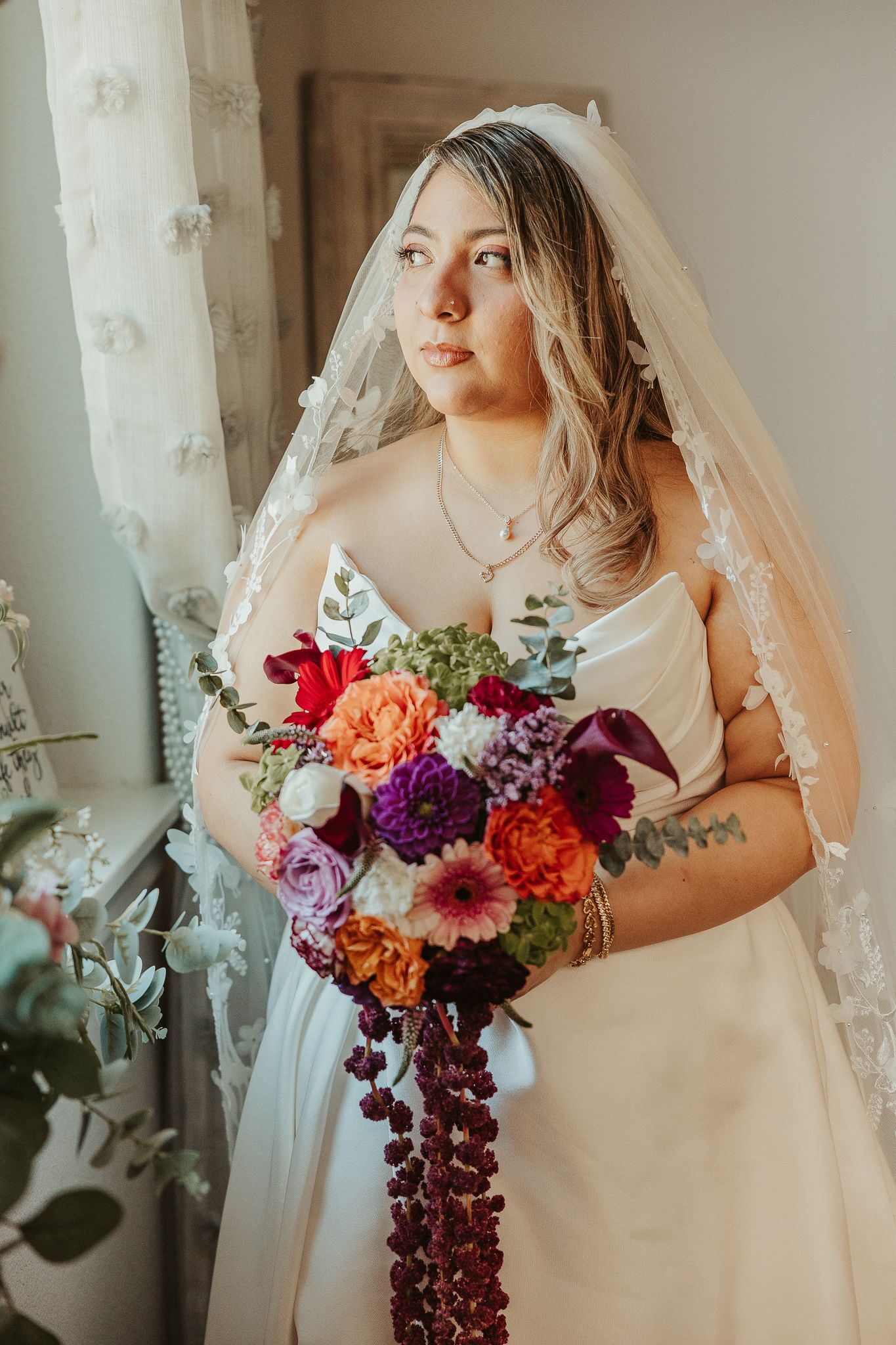 Bride in white gown and lace veil holds colorful bouquet with purple, red, and orange flowers near a window.