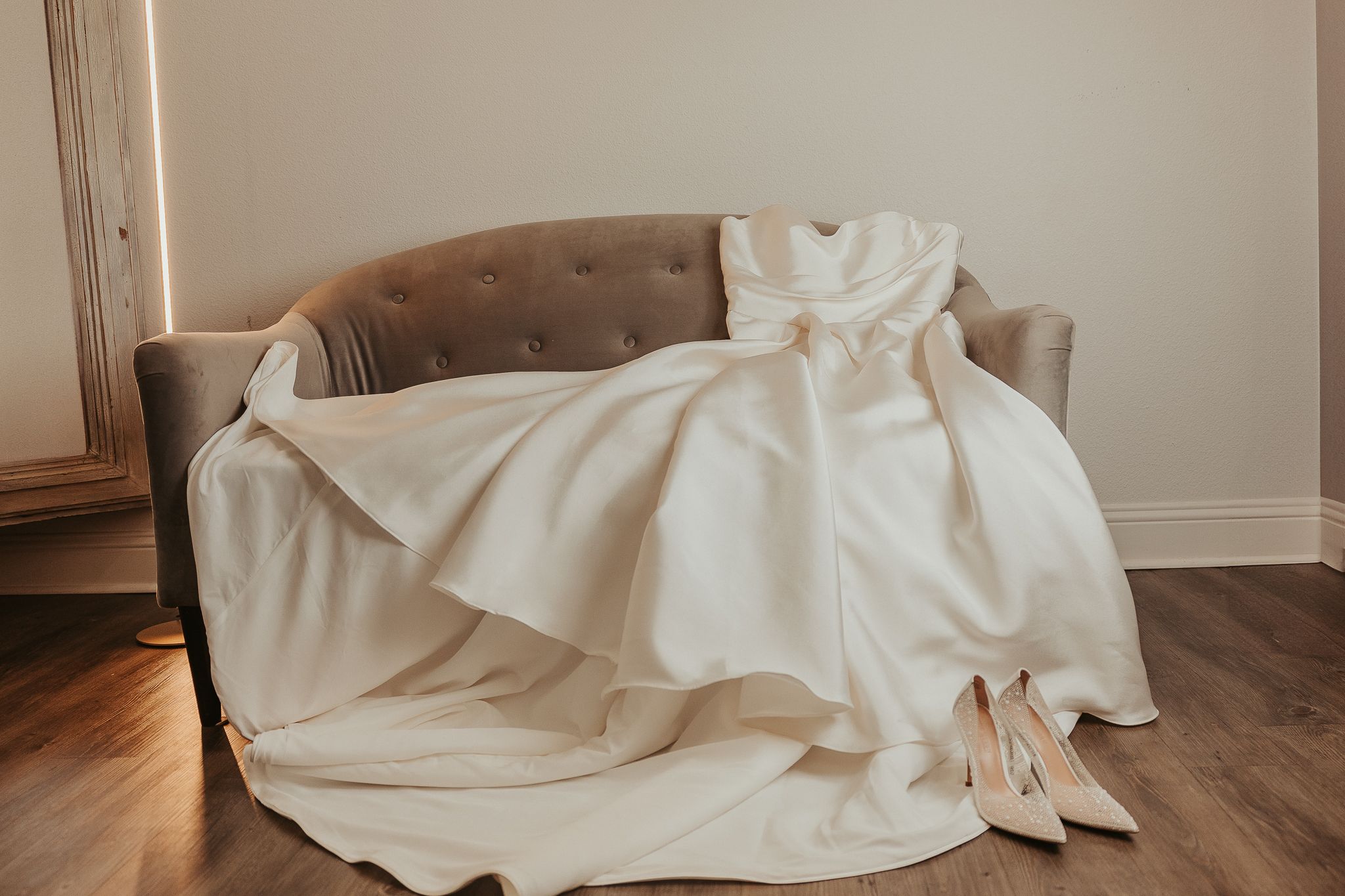 Elegant ivory wedding gown draped over a velvet sofa with matching bridal heels on hardwood floor.