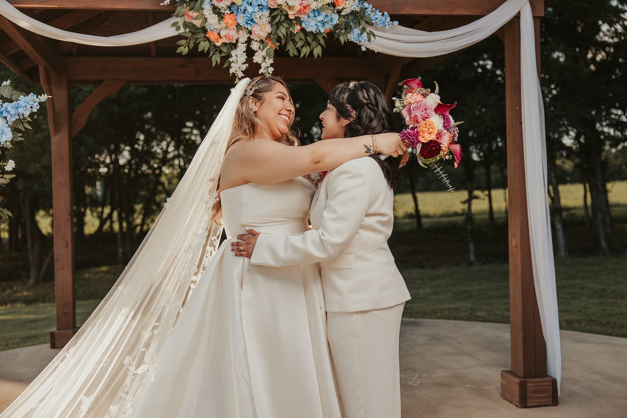 Two brides embrace and laugh joyfully at outdoor wedding ceremony under floral arch, one in white gown with veil, other in white suit.