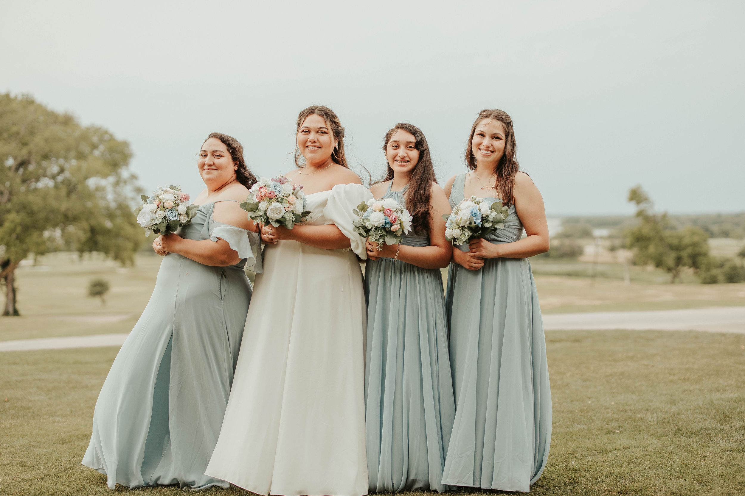 Bridesmaids in flowing sage green dresses hold white bouquets while standing together outdoors.