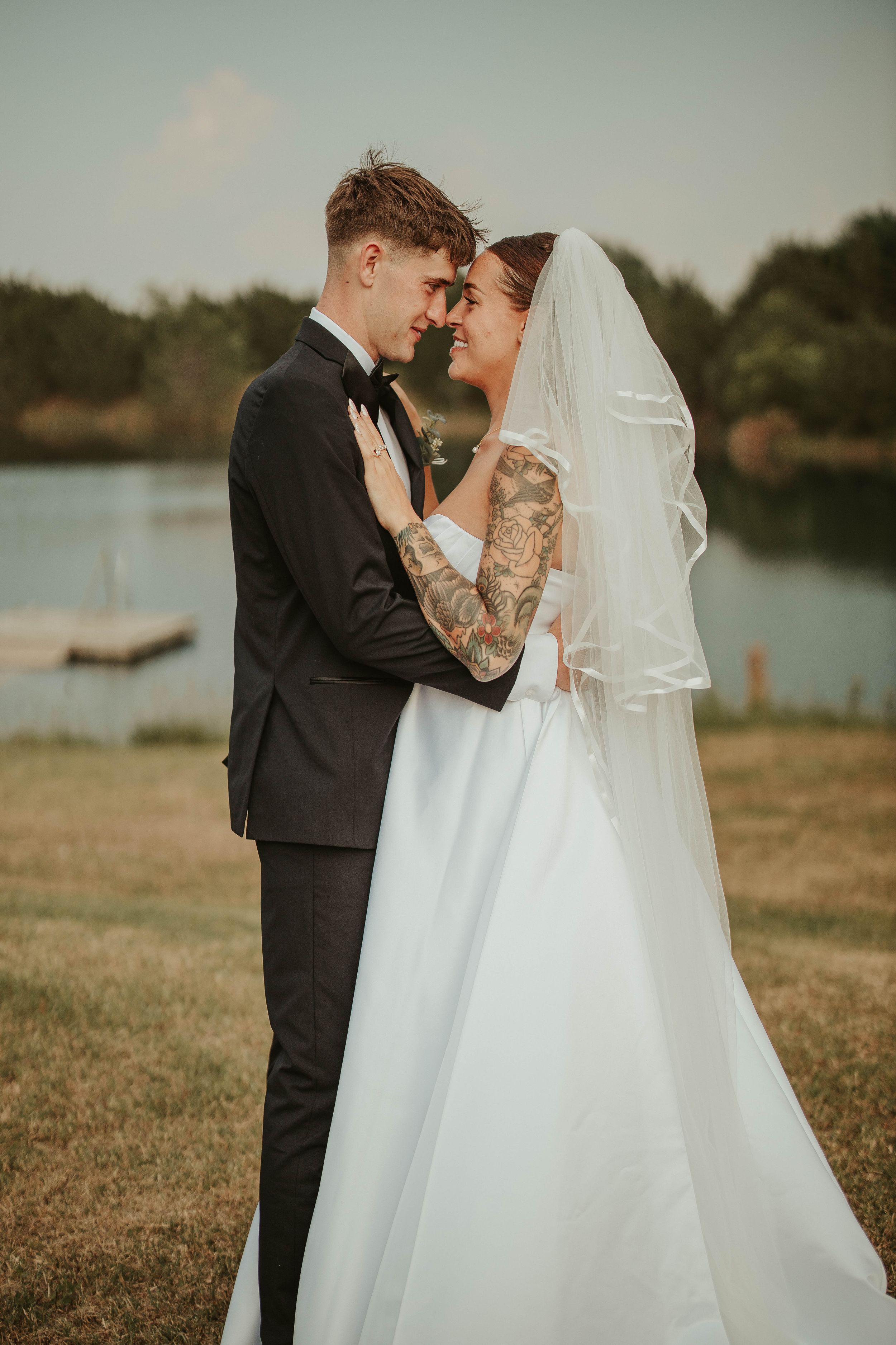 A romantic wedding photo showing a couple embracing outdoors near a lake during golden hour.