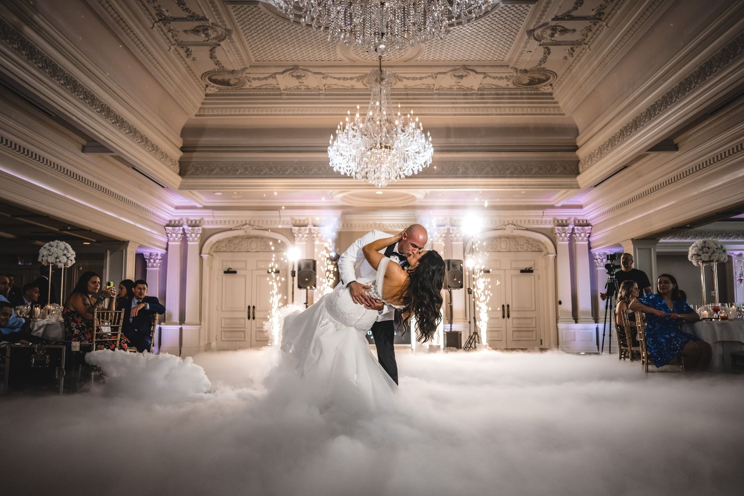 A couple dances in an elegant ballroom with crystal chandeliers and ornate architectural details.