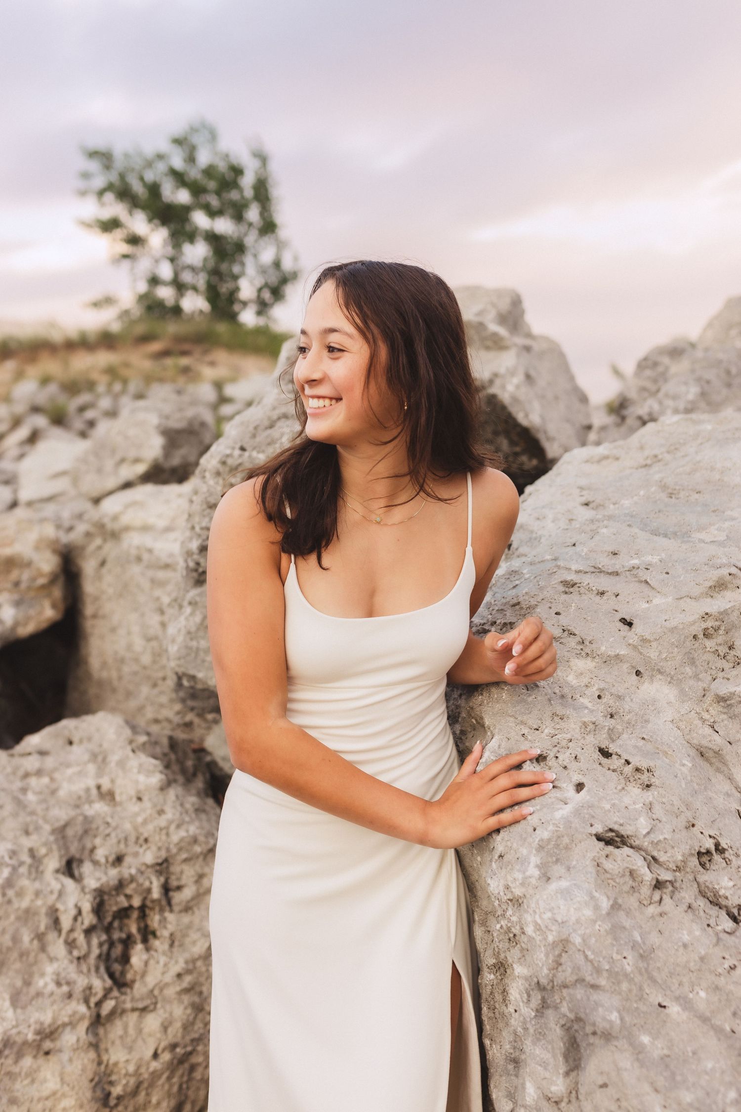 A figure in a flowing white dress poses against large limestone rocks at sunset with soft lighting and windswept hair.