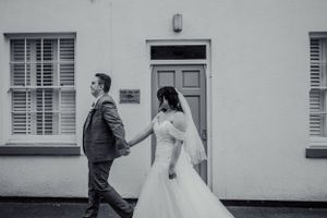 Bride and groom in formal attire stand back to back against white building exterior.