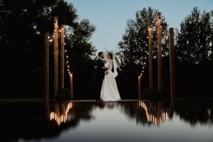 Bride and groom silhouetted against illuminated pillars reflecting in still water at night.