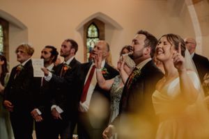 Wedding guests sing together during church ceremony with stained glass windows visible.