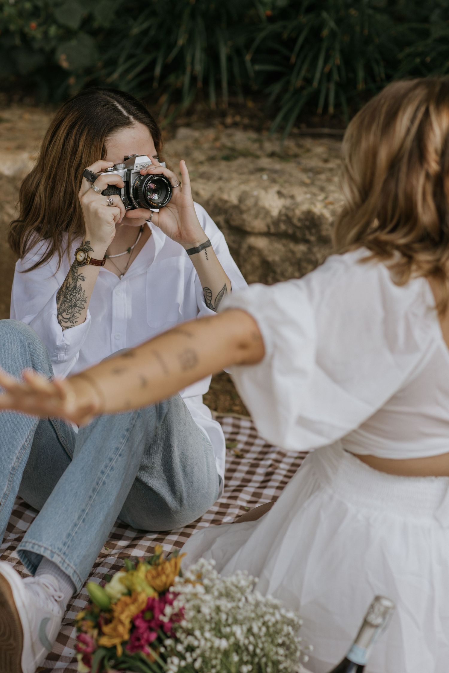 Two friends sitting on picnic blanket sharing a drink from stylish glass cups on a sunny afternoon.