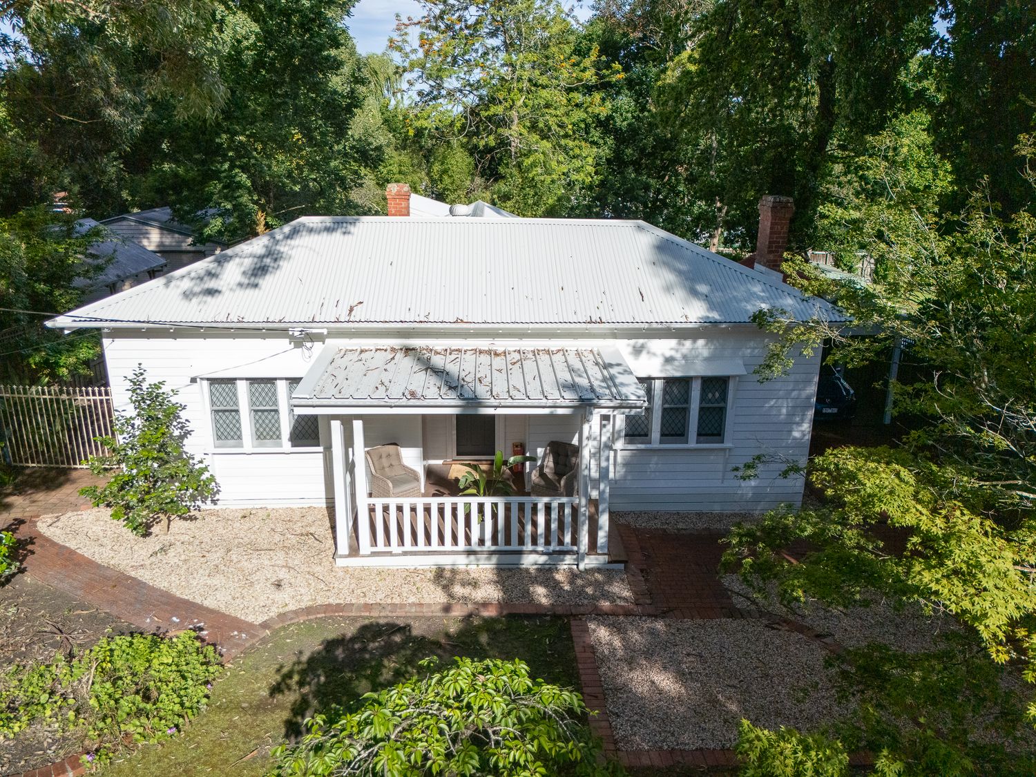 Classic white weatherboard cottage with tin roof, timber veranda, and chimney nestled among mature leafy trees.