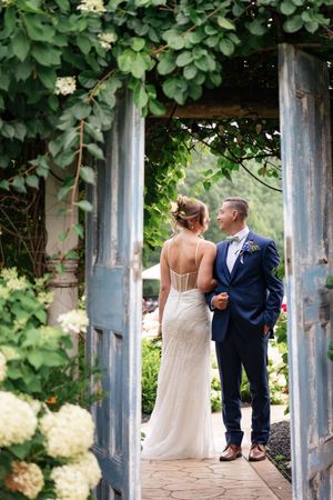 Bride and groom standing in rustic blue doorway framed by lush greenery and white hydrangeas at The Promise Gardens of the Adirondacks.