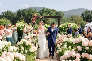 Newlyweds walking back down aisle lined with white hydrangeas as guests applaud outdoors.
