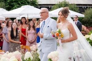 Bride walking down aisle with father carrying colorful bouquet among white floral arrangements.
