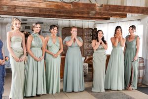 Six bridesmaids in sage green gowns reacting with joy and surprise in rustic barn setting at The Promise Gardens of the Adirondacks.