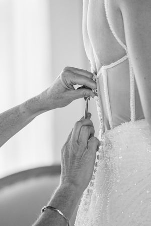Black and white photo of hands fastening buttons on back of beaded wedding dress.