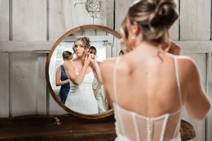 Bride adjusting earring while reflected in round mirror with bridesmaid assisting in rustic room at The Promise Gardens of the Adirondacks.