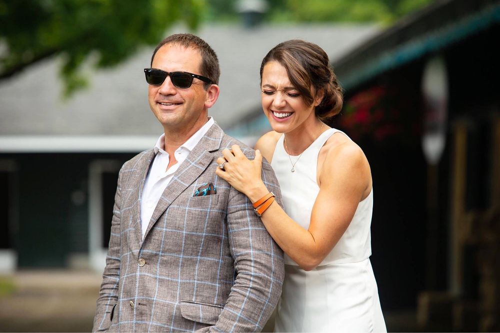 Cherie DeVaux in white gown laughs and David Ingordo in plaid blazer smiles in sunglasses outside at Fasig-Tipton in Saratoga Springs, NY.