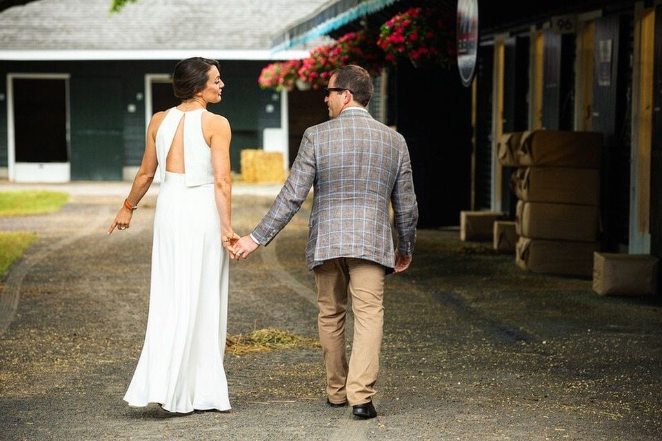Cherie DeVaux in white gown and David Ingordo in plaid blazer walk hand in hand outside at Fasig-Tipton in Saratoga Springs, NY.