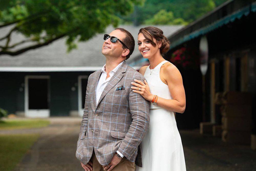 Cherie DeVaux in white gown and David Ingordo in plaid blazer smile and pose outside at Fasig-Tipton in Saratoga Springs, NY during wedding.