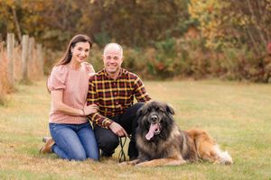 Couple smiling with large Leonberger dog outdoors in autumn park setting.