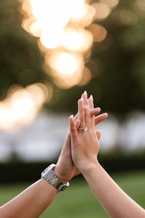 Close-up of couple raising hands together showing diamond engagement ring against golden sunset bokeh background The Sagamore Resort.