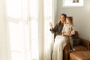 A smiling mother waves out a sunlit window while holding her young daughter on a leather armchair.