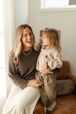 A laughing mother in a brown sweater sits with her toddler daughter on a leather chair by a bright window.