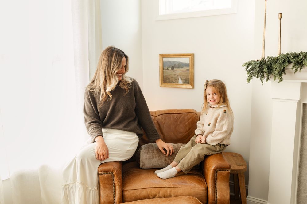 Mother and young daughter sitting together on brown leather armchair in bright cozy living room.