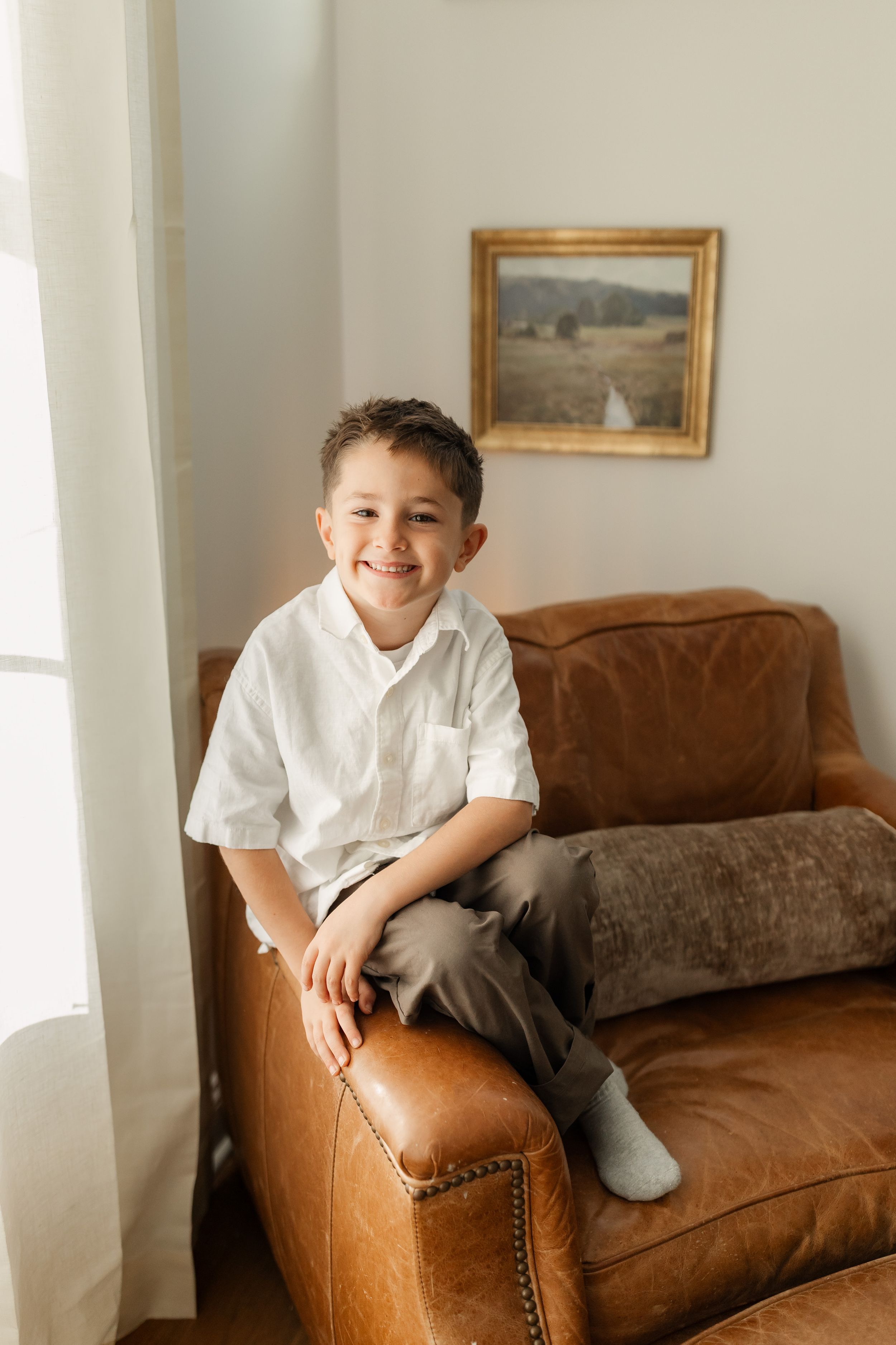 Smiling young boy in white shirt sitting on brown leather armchair in bright home interior.