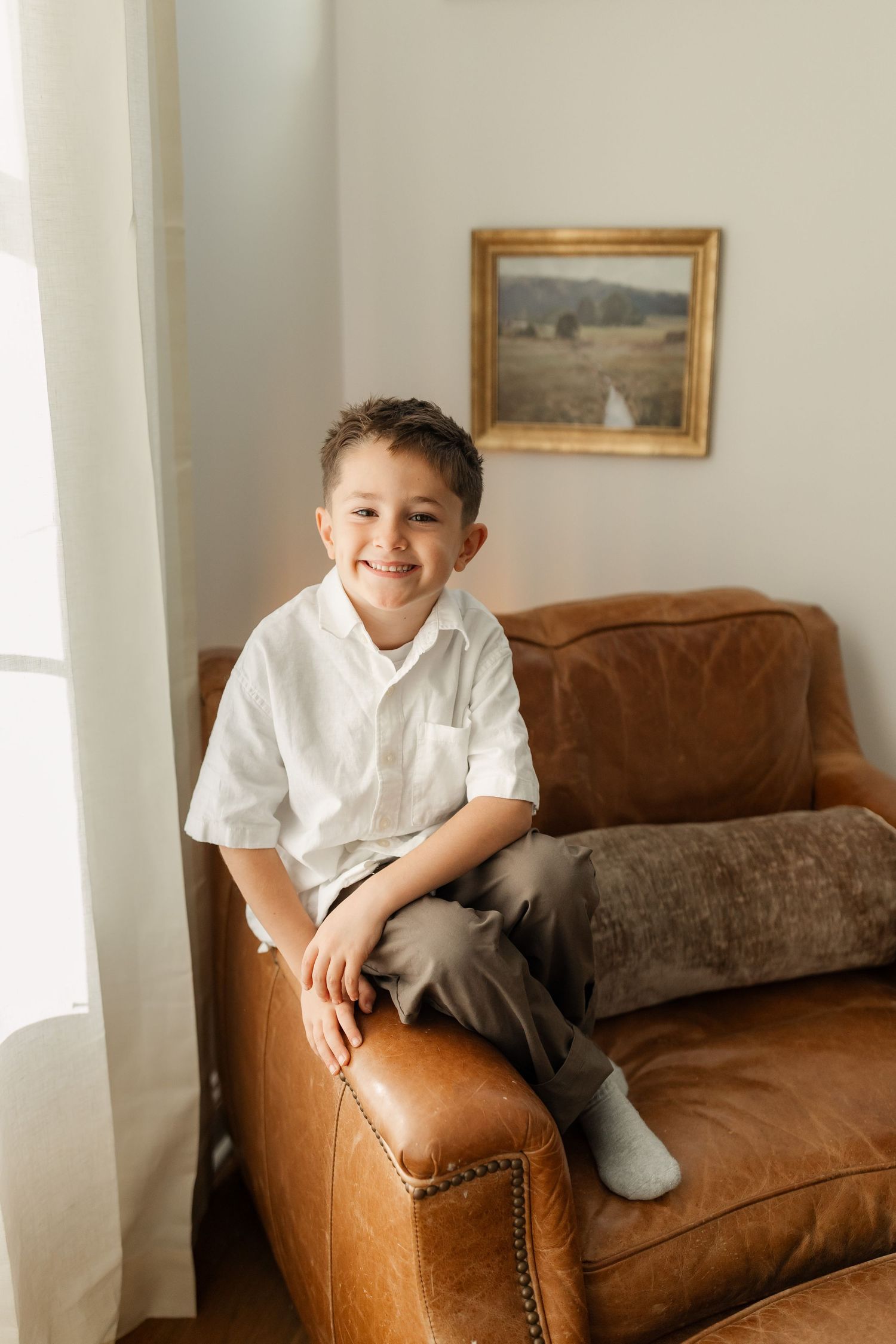 Smiling young boy in white shirt sitting on brown leather armchair in bright home interior.