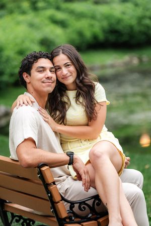 Couple sitting on ornate park bench, woman in yellow dress embracing man, green pond in background.