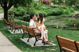 Engaged couple sitting on park bench facing each other near scenic pond surrounded by greenery.