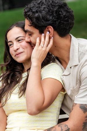 Newly engaged couple embracing, woman smiling showing engagement ring while man kisses her cheek outdoors.