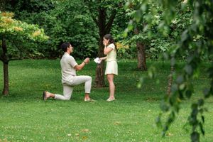 Man proposing on one knee to woman in yellow dress on green lawn in park setting.