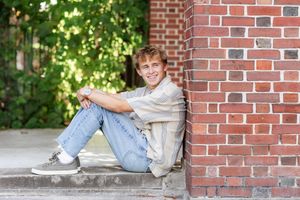 Smiling teenage boy in casual outfit sitting on steps leaning against a red brick wall during Senior Portraits at Saratoga Spa State Park .