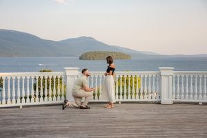 Man proposing on bended knee to woman on scenic waterfront deck with mountain island views at The Sagamore Resort on Lake George, NY.