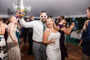Bride and groom dancing at reception wearing novelty glasses, holding drinks under white tent.