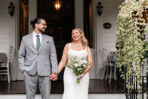 Bride and groom holding hands smiling on white venue porch, bride holding greenery bouquet, groom in gray suit.